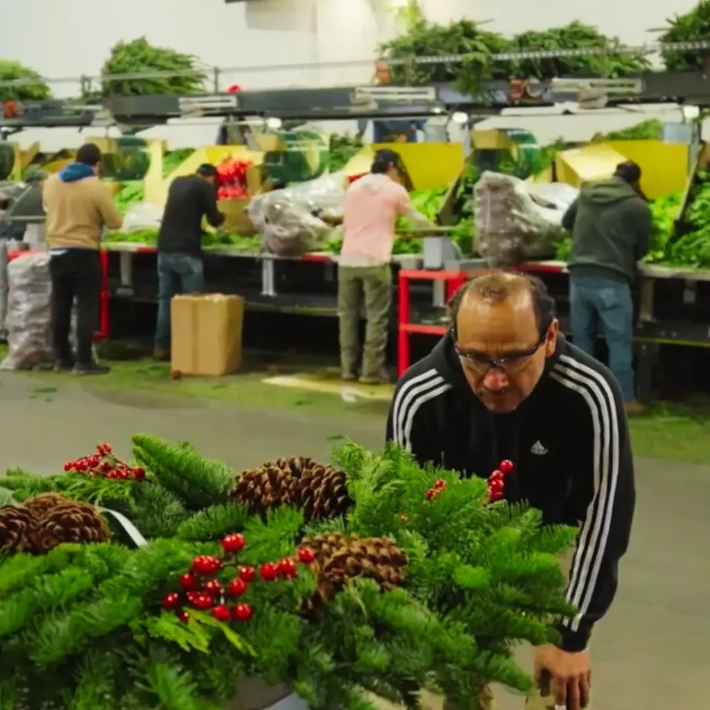 Seasonal worker in safety glasses finishes a Christmas wreath with pine cones and berries while others work at assembly stations behind him