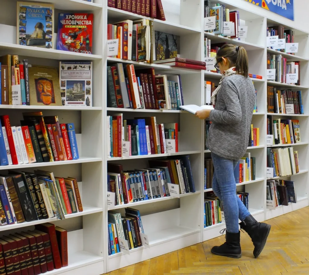 Student reading a book in front of tall white library shelves with labeled sections