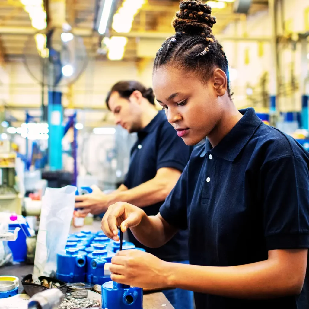 Two production workers assemble blue machined parts at a workbench stocked with components