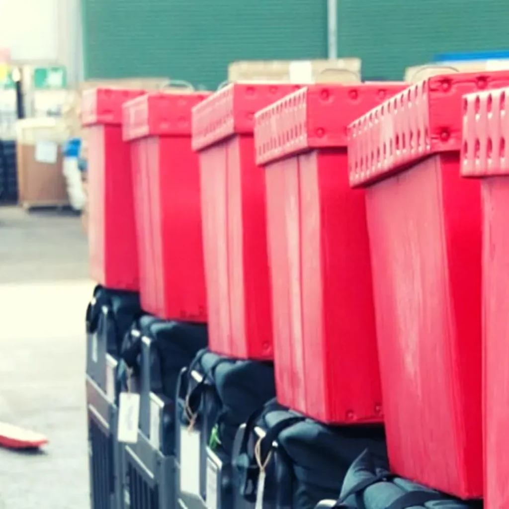 Row of red returnable plastic totes with lids stacked on carts in a warehouse, each with a tag